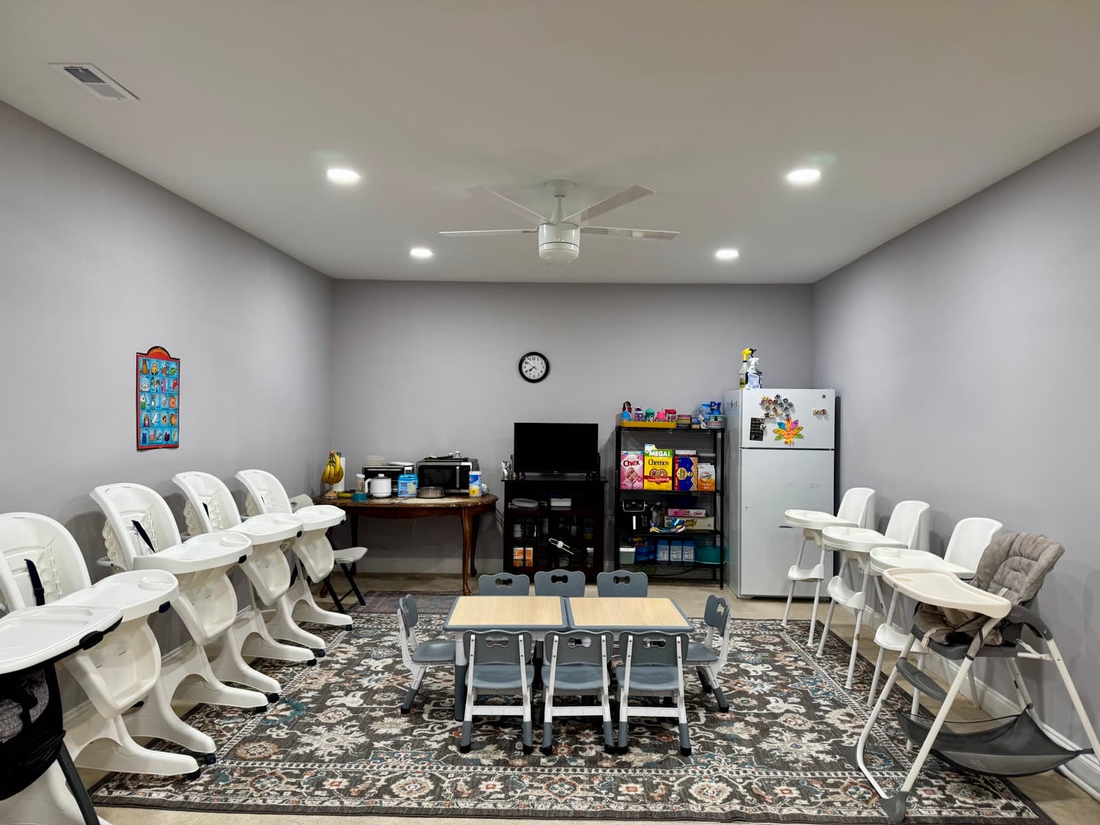 Mealtime and activity room with a row of infant high chairs on either side and a low table with toddler-sized chairs in the center.