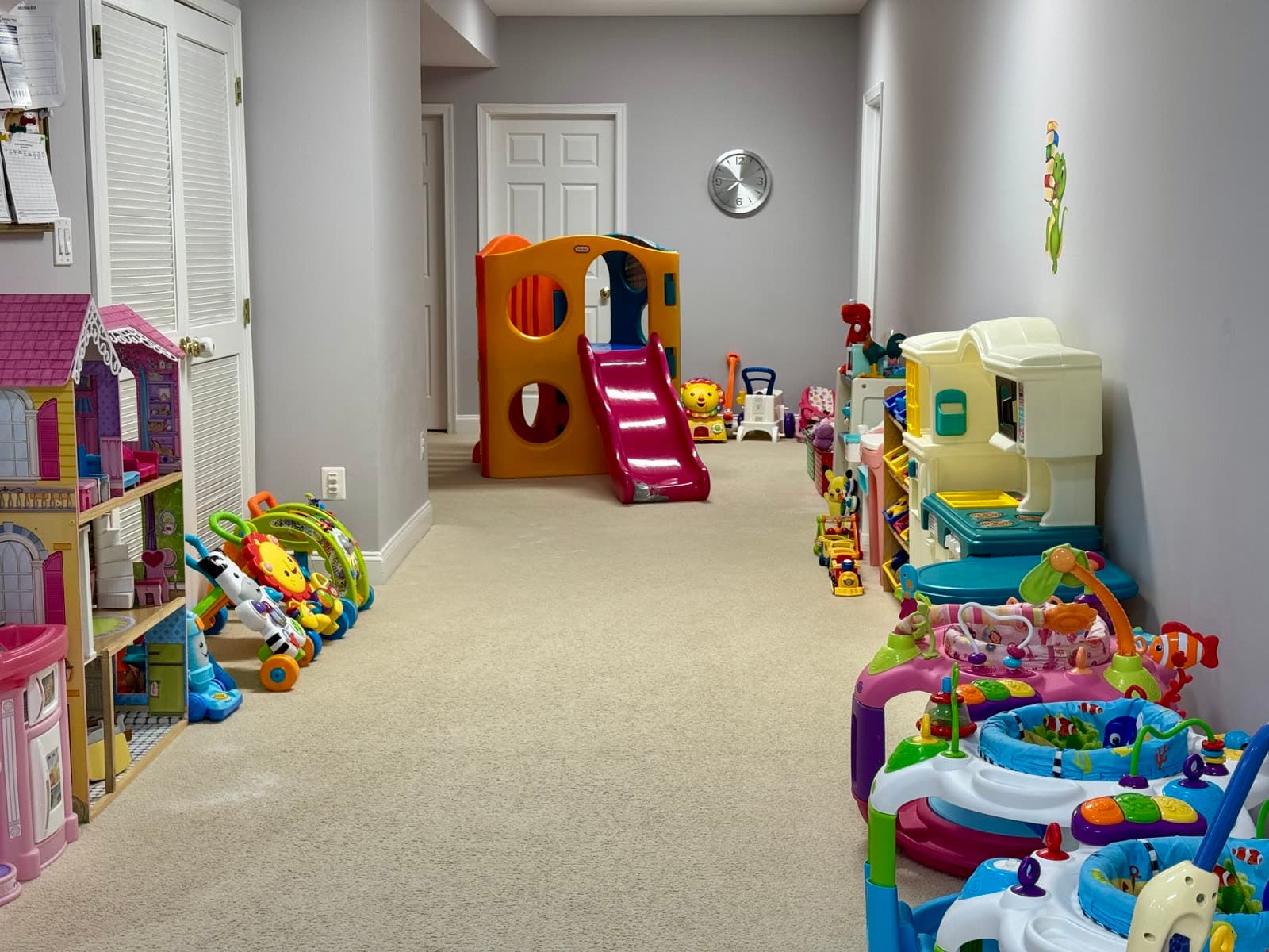 Hallway play space lined with a play kitchen, dollhouse, activity tables, and a small climbing slide at the end.