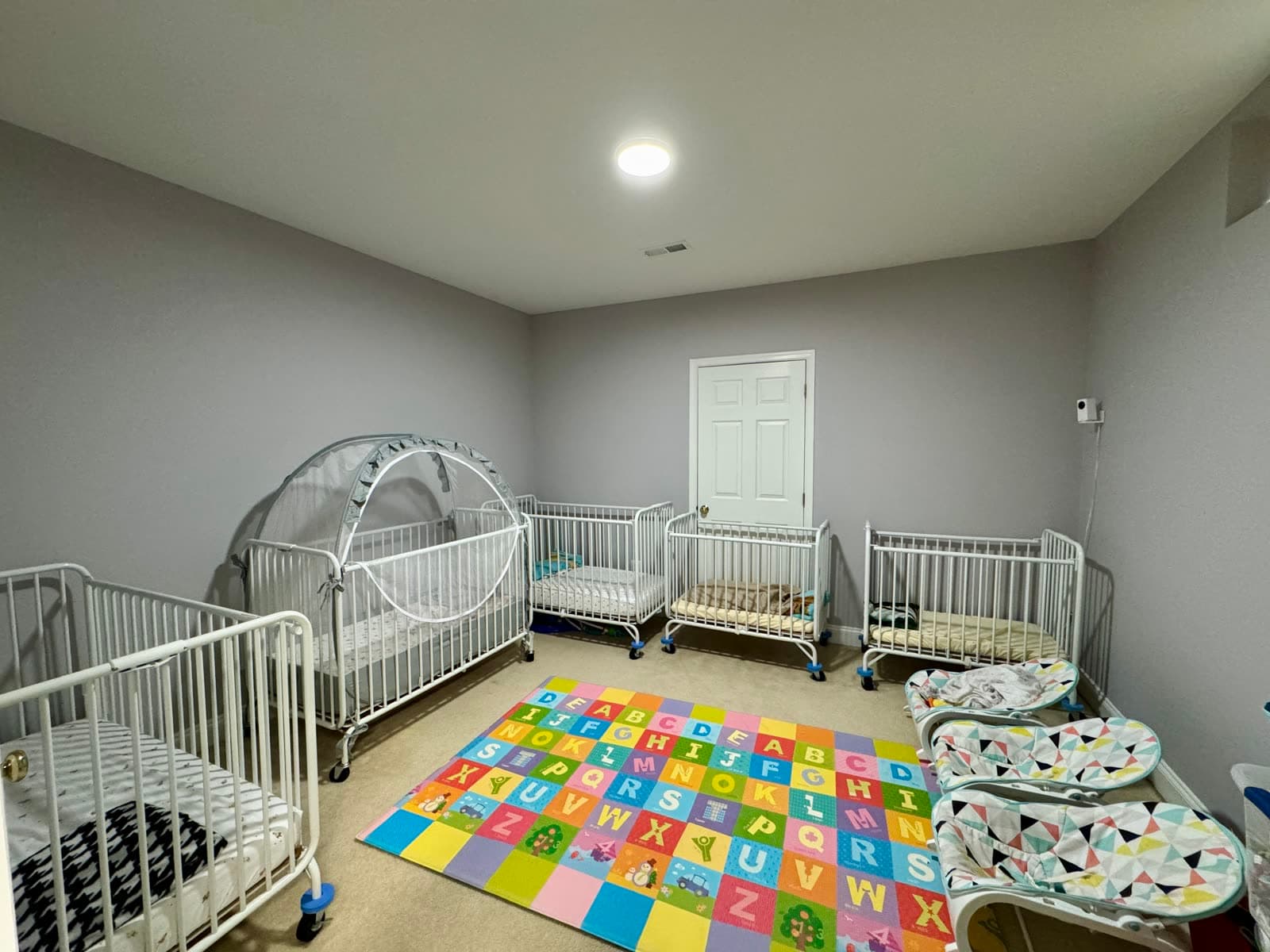 Nap room with several portable cribs arranged around a colorful alphabet floor mat.