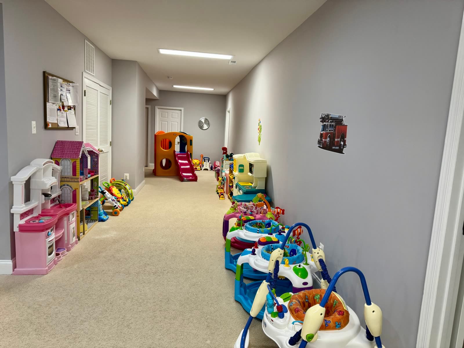 Hallway lined with toy shelves, a play kitchen, and bounce/activity seats for babies.
